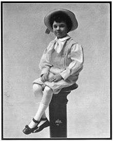 A black-and-white photographed of a neatly-dressed young boy in a hat, sitting on a pedestal