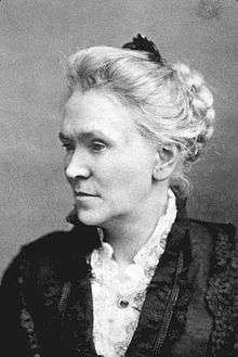 A faded monochrome photograph portrait of a woman turned to the left, facing further left, wearing a dark coat closed with a floral brooch, over a white blouse with a lacy collar, the woman's blond hair held up in a bun on the back of her head, with a dark hair comb standing vertically in the bun