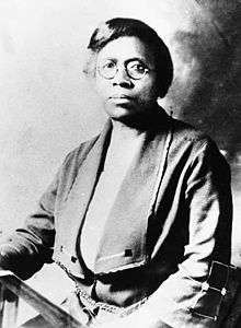 An African American female with short dark hair, round eyeglasses, and a solemn expression sitting down and facing the camera.