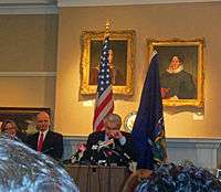 A man at a lectern with his hand to his face. Behind him are the United States and New York flags and a wall with portrait paintings