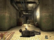 A video game screenshot of a man leaning on the floor while holding a gun in each hand and firing them at another man down a hallway.