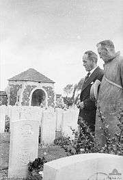 Photograph of two men in suits standing over to the left gazing at a grave marker. Several other graves and a structure are in the background.