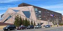 An angular reddish-gray building stands before a brown building on a clear day.