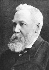 A head and shoulders view of an elderly man with white hair and a large, bushy beard. He is wearing a dark jacket and tie and a white shirt