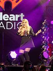 A young long-haired blonde woman singing into a microphone onstage. She wears a black skirt and black Bad Gal jacket while pink stage lighting shines upon her. An iHeartRadio logo is her backdrop.