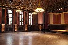 A ballroom with hardwood floors, and a ceiling decorated with chandeliers and octagon-shaped tiles. The wall on the left is decorated with dark wood panels, red walls, and windows. On the right is a small stage with a piano.