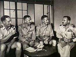 A group of four senior military officers sitting down together conversing. There is a small table in front of them with a book on it.
