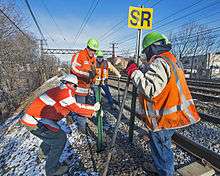Four men in reflectorized orange vests and neon green helmets standing next to a set of four railroad tracks installing a signpost next to the track. One is holding a sign with the letters "SR" in black on yellow. The ground, covered with a thin snow, slopes steeply to the left and an overhead gantry for the catenary is in the background.