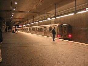 A train is waiting on a track in a subway station. Passengers are standing on the platform.