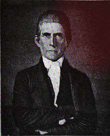 A man with slicked-back, dark hair wearing a white shirt and dark jacket