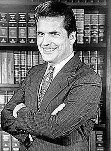Greyscale photo of a man in a suit with arms crossed, a smile, and a tie. Background has bookshelves of law books.
