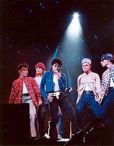 Under a spotlight, a man sings into a microphone. He is surrounded by four male dancers dressed up to look aggressive. The man himself wears black pants held up by a white sash, as well as a white T-shirt, and an opened blue shirt, which is tucked into his pants.