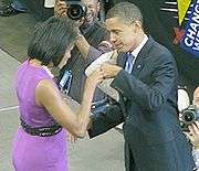 The Obamas face each other and bump fists on stage. She wears a purple dress and he wears a dark suit. Several signs read "CHANGE WE CAN BELIEVE IN" and several photographers take photos.