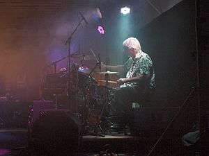 A man with gray hair drums behind his drum-set in a faintly lit room.
