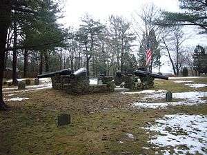 A monument featuring four black cannon barrels mounted on a stone wall in the middle of a small cemetery.  The ground is partly covered with snow.  Many trees stand in the background.  The sky is cloudy.