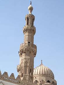 An ornate carved stone minaret, with a carved stone railing around three balconies, the first below its center, the second two thirds the way up, and the third near its top. The tip of the minaret is a large bulb-shaped stone decoration with a small bulb-shaped metal finial. Behind the minaret most of a dome is visible.