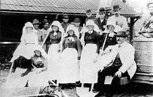 Large group of men in bowler hats and women wearing large bonnets, posing with tools