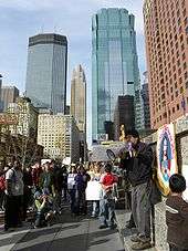 A daytime city scene on a sidewalk with a tall, brick-faced building on the right and glass-covered skyscrapers in the distance in the center. A dark-haired, fairly young man dressed in casual clothes stands next to a drawing of Jesus and speaks through a megaphone. About 25 people of different ages and both genders can be seen watching him, with a few holding signs, one of which says SEIU.
