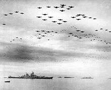 Black and white photo showing a large number of aircraft flying in formation over several World War II-era warships