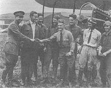 A monochrome photograph of eight variously dressed military airmen standing smiling in front of a biplane, with a tall brigadier general in uniform shaking hands with a short-statured, hatless man wearing a long-sleeved shirt and a tie