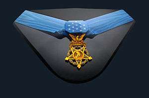 U.S. Army Medal of Honor displayed with a light blue neck ribbon with a gold star shaped medallion hanging from it. The ribbon is similar in shape to a bowtie with 13 white stars in the center of the ribbon.