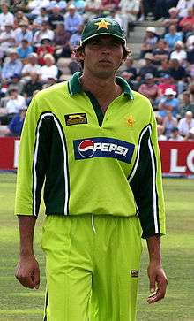 A dark coloured man wearing light green outfit and dark coloured cap. Cricket field and spectators can be seen in background.