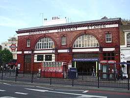 A red-bricked building with a rectangular, white sign reading "MORNINGTON CRESCENT STATION" in black letters all under a light blue sky
