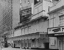 Daytime. A plain theatre front is seen from the sidewalk, at an angle, with an electrical sign above showing the theatre's name. No play is advertised. The cornice is dirty and in disrepair. Next door (at right), part of another theatre is shown, with a play advertised on its marquee, and two "Post No Bills" signs at eye level. The left of the image shows part of a large hotel, with three people near the entrance.