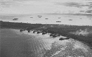 A peninsula with eight ships beached on the shore in the foreground and over eleven ships anchored off the opposite shore. Smoke is rising from the peninsula.
