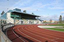 A brown running track lies within a small, open stadium below a blue sky.