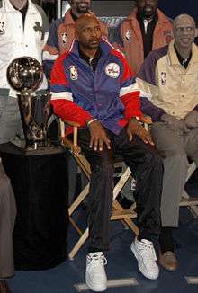 A man, wearing a black shirt and blue-red jacket with the logo NBA on it, is sitting on a chair while posing for a photo. He is surrounded by four other men, while a gold trophy is placed on his right.