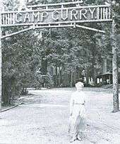 Woman in a long dress in front of a sign across a road. Wooden letters read "Camp Curry".