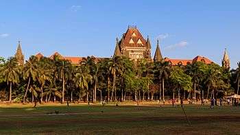 A brown building with a central tower and sloping roofs surrounded by trees. A grassy ground and a coconut tree are in front of it.
