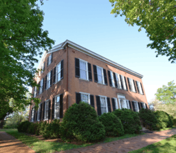 A two-story, brick home with thirteen windows with black shutters and several bushes growing in front