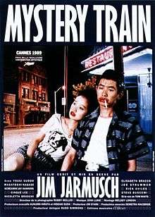 A young Japanese couple are sitting on a dimly-lit street outside a restaurant with a neon sign. The woman is looking off camera and resting against the man, who is downcast with a cigarette in his mouth; both are wearing bright red lipstick. Superimposed above the couple is the title MYSTERY TRAIN, with credits in French at the bottom of the image.