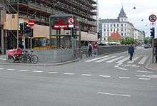 A street intersection. In a traffic island is an elevator marked "Nørreport" and beside it is the top of an escalator