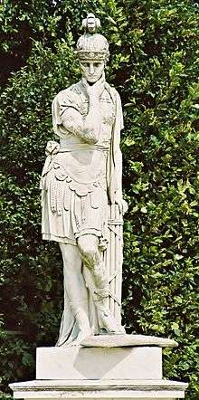 A white statue of a man in front of a green hedge. The statue was sculpted in the fashion of the Roman army, wearing the helmet and livery of the army. The statue has one arm raised up to its chin and the other behind it, resting on another piece of the sculpture.