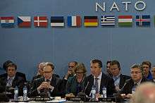 An older man in a suit sits, speaking at a round table with dozens of other men and women. Above them is the word NATO and nine European flags.