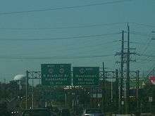 A four lane divided highway running through a business district with two overhead green signs. The left sign reads west Route 38 to Route 41 Ben Franklin Bridge Haddonfield 1/2 mile while the right sign reads Route 38 east Moorestown Mount Holly keep right