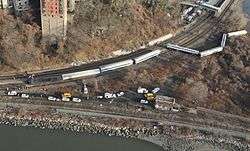 An aerial view of a wooded area between two watery areas in late autumn, with two railroad tracks forking to the top near the left and one following the water's edge. Several silvery train cars and a locomotive are lying on their sides off the tracks to the top; there are many parked yellow and white trucks and other vehicles between the two tracks.