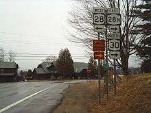 A pair of highways intersect in front of a pair of cabins in a rural area. The highway in the foreground ends here, forcing drivers to turn left or right. A nearby sign assembly indicates that NY 28 west is to the left while NY 28N east and NY 30 north are to the right.
