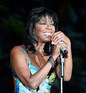 A woman in her late fifties. She wears a blue sleeveless top and is holding a microphone and smiling.