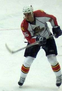 An ice hockey player has his stick in the air trying to hi an unseen puck. He is looking at his stick with a focused look on his face. He wears a "traditional" white Panthers jersey with the number 16 on the sleeves.