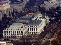 An aerial view of a white stone neoclassical building. There is a large brown dome towards the front and center of the building where the entrance is.