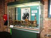 A glass display case containing a large brass model of a steam locomotive. At the back of the case is a black and white photograph of a man.