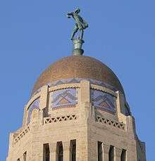 Gold-colored dome atop octagonal stone tower; stylized thunderbirds on sides of tower just below dome; bronze statue of someone sowing seed by hand on top
