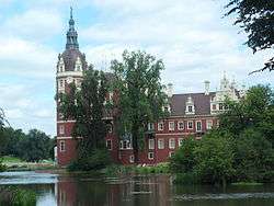 A red, ornate neo-gothic castle in a park-like location, the main tower of the castle is located to the left and topped with an ornate round dome and spire