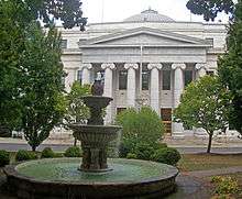 The courthouse seen with a fountain in the foreground and tree branches on the sides. Part of the dome is visible on top.