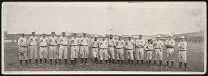A single row of men in white baseball uniforms with high socks and white baseball caps standing on a baseball field; their uniforms read "NY" across the chest.