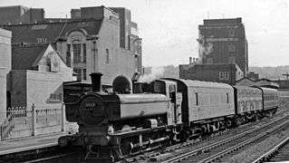 A pannier tank locomotive pulling a train of three wagons through a station. The coaches are a parcel van, a goods wagon, and a passenger coach.